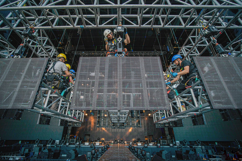 Rigging crew in hard hats and harnesses installing rental LED wall panels on a truss grid above a stage